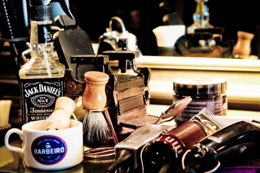 ParaTodosVerem: Em uma bancada de barbearia, diversos itens estão organizados próximos a um espelho iluminado. À esquerda, uma garrafa de Jack Daniel's é usada como decoração ao lado de uma caneca branca com o logotipo “Sr. Barbeiro”, contendo um pincel de barbear com cabo claro. Há também outro pincel de barbear apoiado na superfície. Ao fundo, destacam-se máquinas de cortar cabelo antigas em estilo retrô(Foto: ETHI ARCANJO)