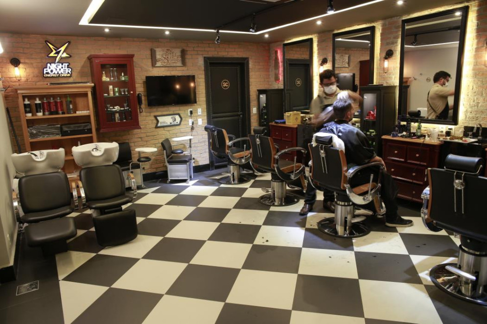ParaTodosVerem: Interior de uma barbearia sofisticada com piso quadriculado em preto e branco. À direita, um barbeiro de máscara realiza um corte de cabelo em um cliente sentado em uma das cadeiras giratórias de couro preto com detalhes em madeira. À esquerda, há lavatórios e cadeiras de espera. As paredes são de tijolos aparentes, com móveis de madeira escura, prateleiras com produtos de beleza e uma televisão fixada na parede(Foto: JÚLIO CAESAR)