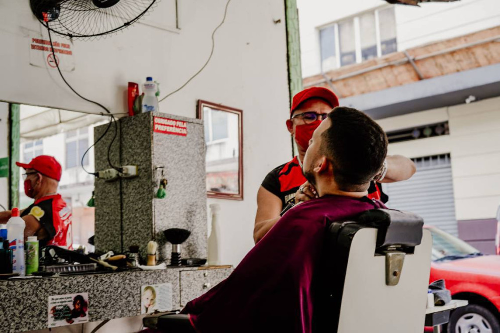 ParaTodosVerem: Em uma barbearia simples e tradicional, um barbeiro de máscara vermelha, boné e camisa preta com detalhes vermelhos faz o acabamento da barba de um cliente, que está sentado em uma cadeira reclinável com capa roxa(Foto: JÚLIO CAESAR)
