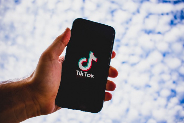 Especialistas debatem o papel dos algoritmos na disseminação de conteúdos violentos. Na imagem, uma mão segura um celular cuja tela mostra o logo do TikTok e ao fundo, um céu azul encoberto por nuvens(Foto: Reprodução/Instagram/Kon Karampelas )