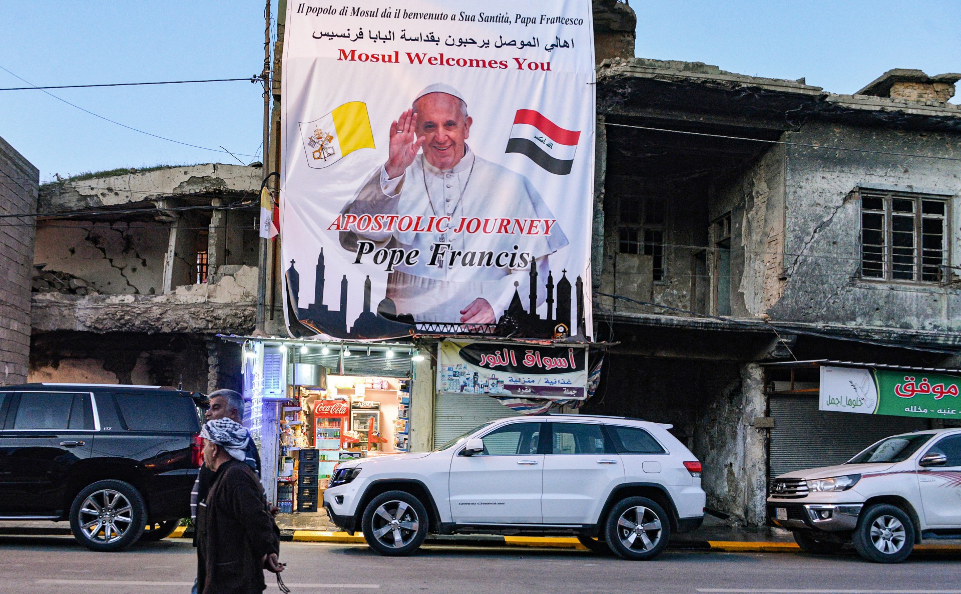 Homens passam por um pôster de boas-vindas ao Papa Francisco pendurado acima de um prédio em ruínas na cidade velha de Mosul, no norte do Iraque (anteriormente devastada por militantes do grupo do Estado Islâmico) em 6 de março de 2021 em meio aos preparativos para a visita do pontífice. (Foto de Zaid AL-OBEIDI / AFP) 