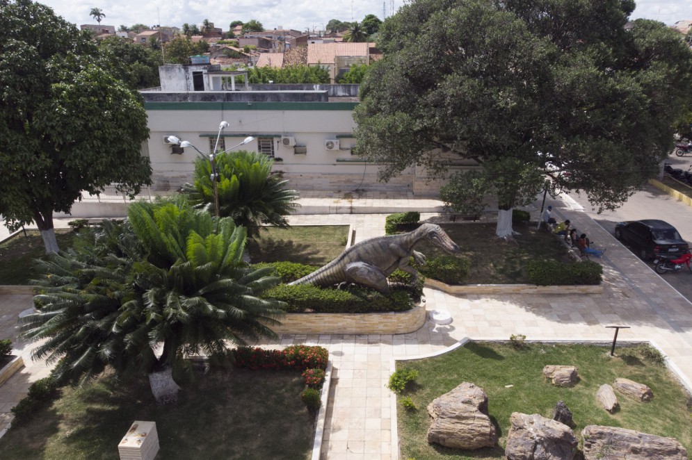 Praça em frente ao Museu de Paleontologia, em Santana do Cariri. No meio, a escultura de um dinossauro. No canto inferior direito, um tronco fóssil de seis metros e cinco toneladas.(Foto: AURÉLIO ALVES)