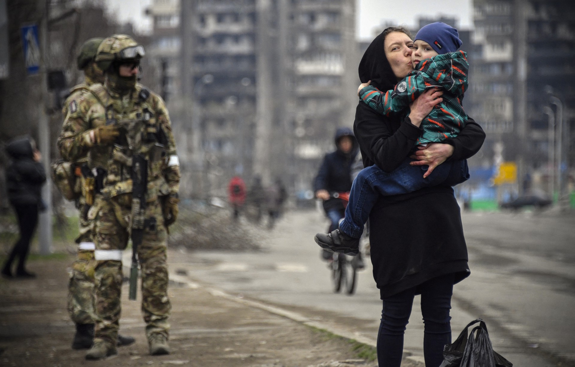 MULHER segura e beija uma criança ao lado de soldados russos em uma rua de Mariupol (Foto: ALEXANDER NEMENOV / AFP)