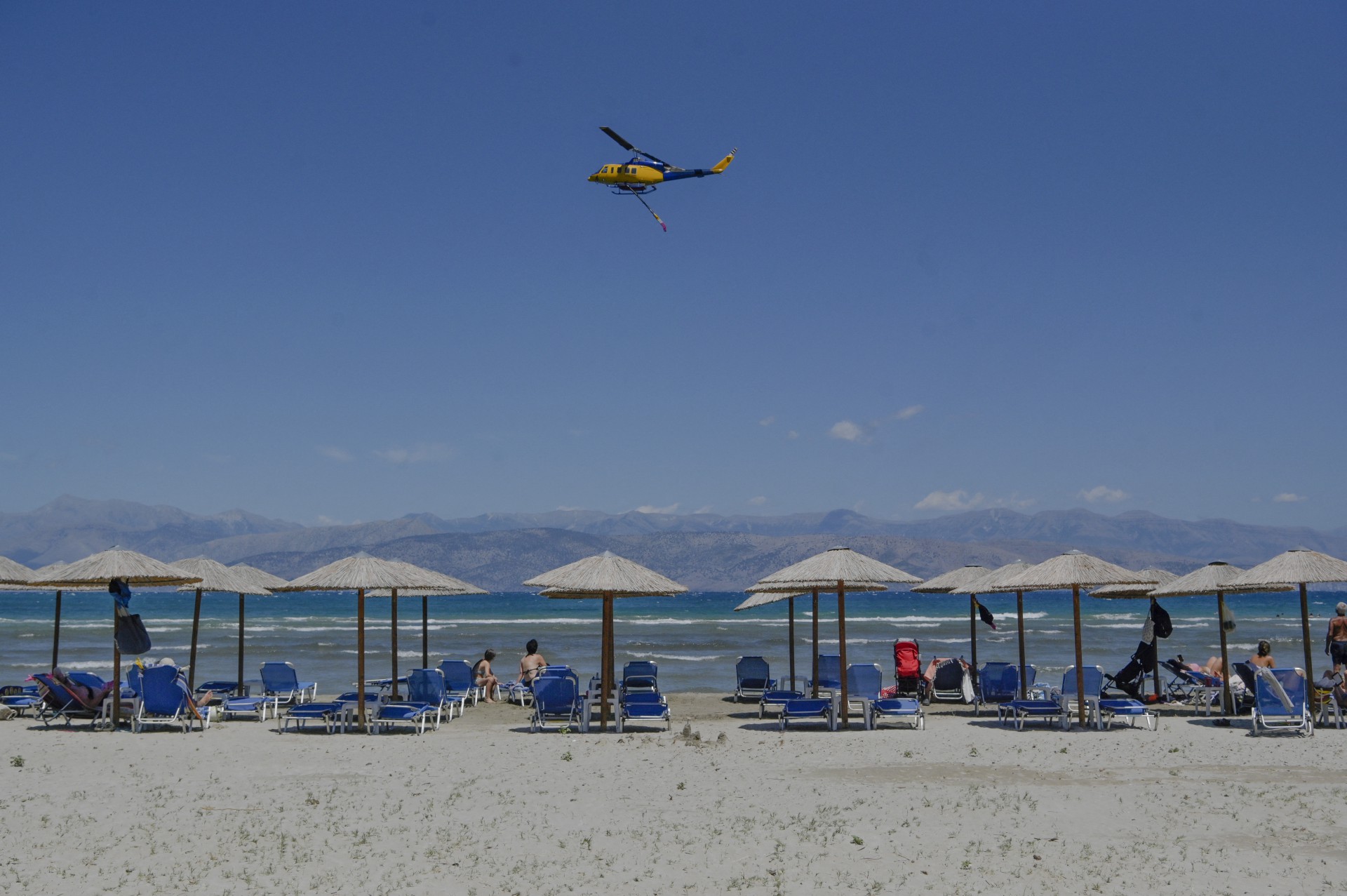 Um helicóptero de combate a incêndios sobrevoa turistas à beira-mar, depois de fazer uma queda d'água em incêndios florestais perto da vila de Loutses, na ilha grega de Corfu (Foto: Armend NIMANI / AFP)