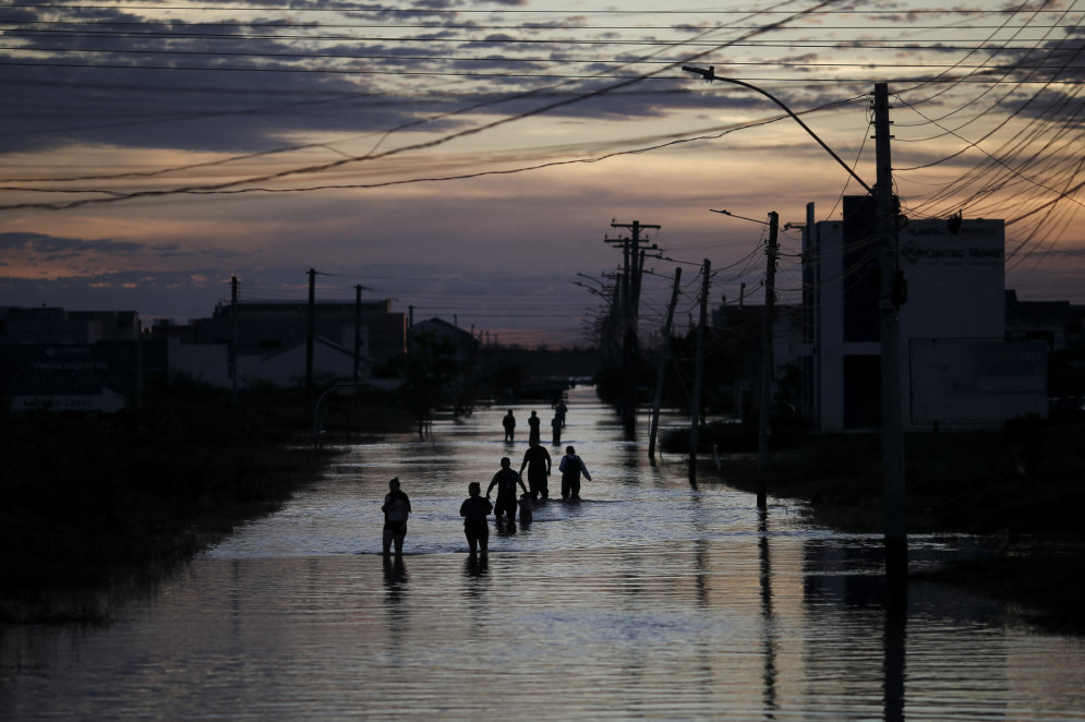 Na imagem, pessoas caminham por uma rua inundada de Eldorado do Sul (RS). "A tragédia das águas, fruto do destempero ambiental do ser humano, traz histórias de vidas e mortes em larga escala", escreve Ana Márcia Diógenes, colunista do OP+