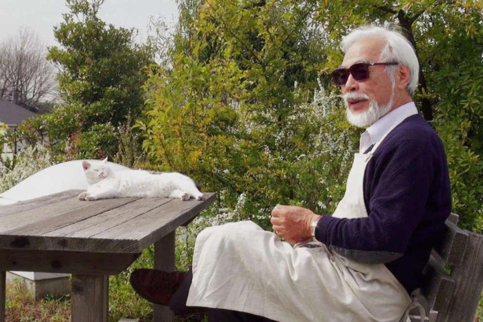 Na imagem, Hayao Miyazaki está sentado em um banco de madeira ao lado de uma mesa de madeira. Ele está usando óculos de sol, um suéter escuro e um avental branco. Em cima da mesa, está um gato branco deitado, relaxando. O fundo mostra um jardim com árvores e plantas verdes(Foto: Cena do documentário Estúdio Ghibli: Reino de Sonhos e Loucura)
