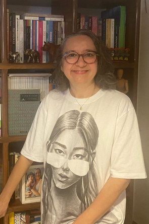 ParaTodosVerem: Maria Erica é uma mulher de pele clara, cabelos grisalhos ondulados, na altura dos ombros, usando óculos de grau e sorrindo levemente(Foto: Reprodução/ Arquivo pessoal )