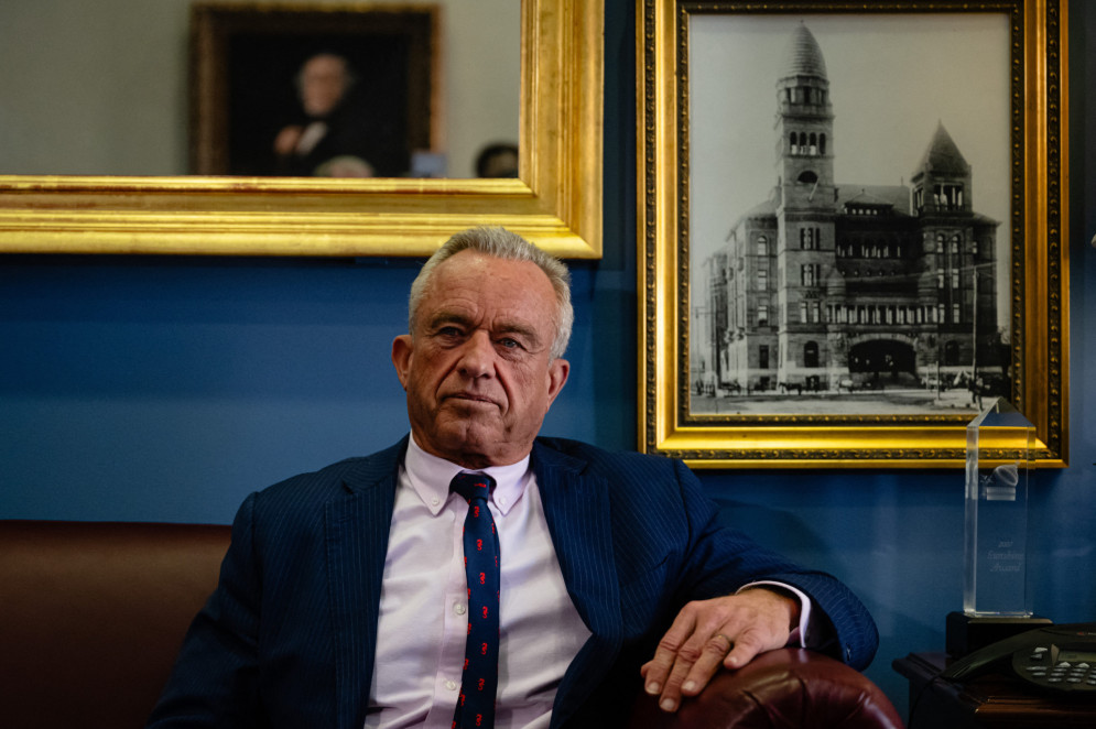 ParaTodosVerem: Secretário da Saúde Robert Kennedy, um homem branco, sem barba e de cabelos grisalhos. Ele está usando um terno e sentado em um sofá de couro. Ele está em um ambiente com uma parede azul, decorada com dois quadros emoldurados: um retrato de um edifício e uma pintura de uma pessoa desfocada ao fundo (Foto: Jon Cherry / Getty Images via AFP)