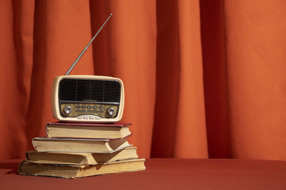 ParaTodosVerem: imagem mostra um antigo aparelho de rádio sobre uma pilha de livros. As peças estão em primeiro plano. Ao fundo, há uma cortina laranja(Foto: Designed by Freepik)