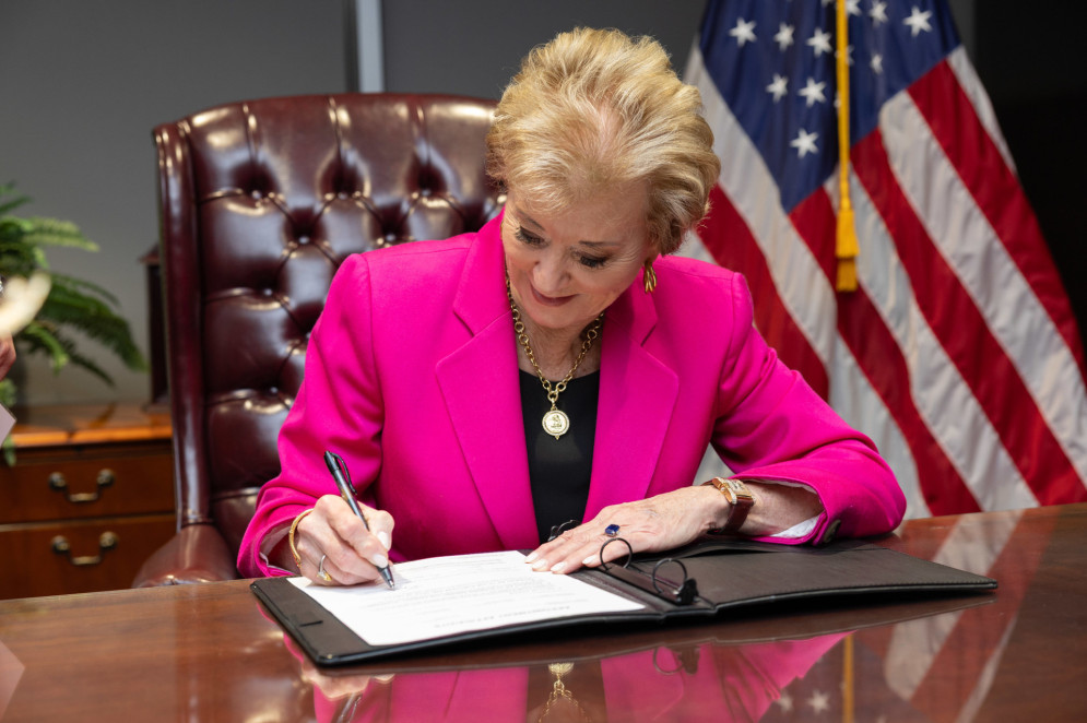 ParaTodosVerem: Fotografia da secretária de educação dos EUA, Linda McMahon. Ela é uma mulher de pele clara e cabelos curtos e loiros, está vestindo uma camiseta preta e um blazer cor-de-rosa. McMahon está sentada em uma poltrona de couro marrom, com uma bandeira dos Estados Unidos ao fundo. Ela está com os olhos voltados para um documento que está sendo assinado por ela(Foto: Paul Wood / Departamento de Educação dos Estados Unidos)