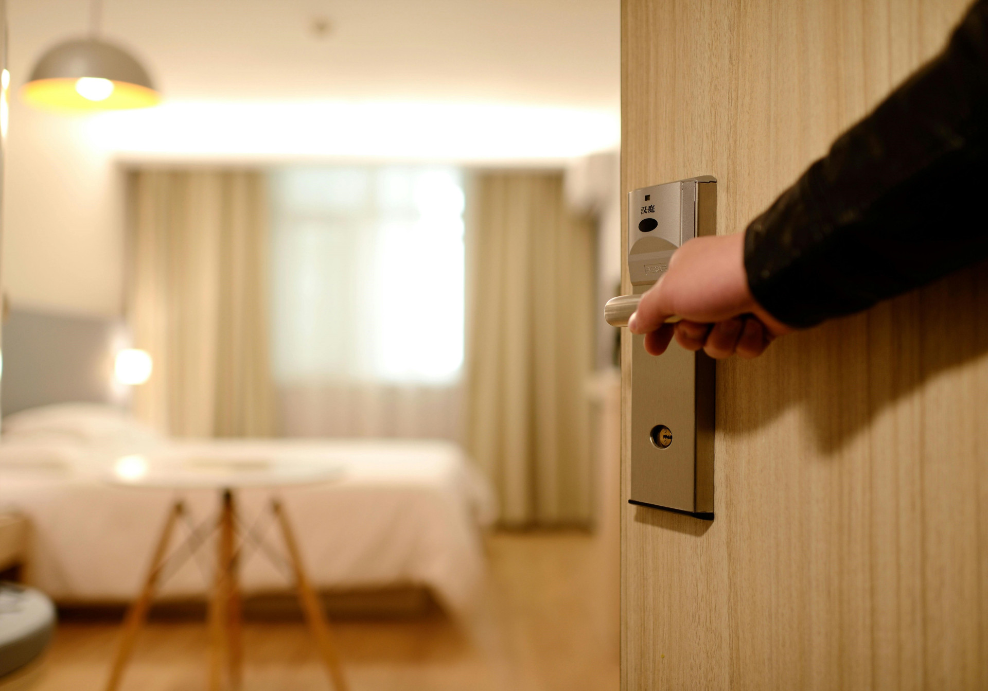 ParaTodosVerem: mão abrindo a porta de um quarto de hotel. Ao fundo, cama de casal, mesa e cortinas claras, em ambiente iluminado. (Foto: Pexels/Pixabay)