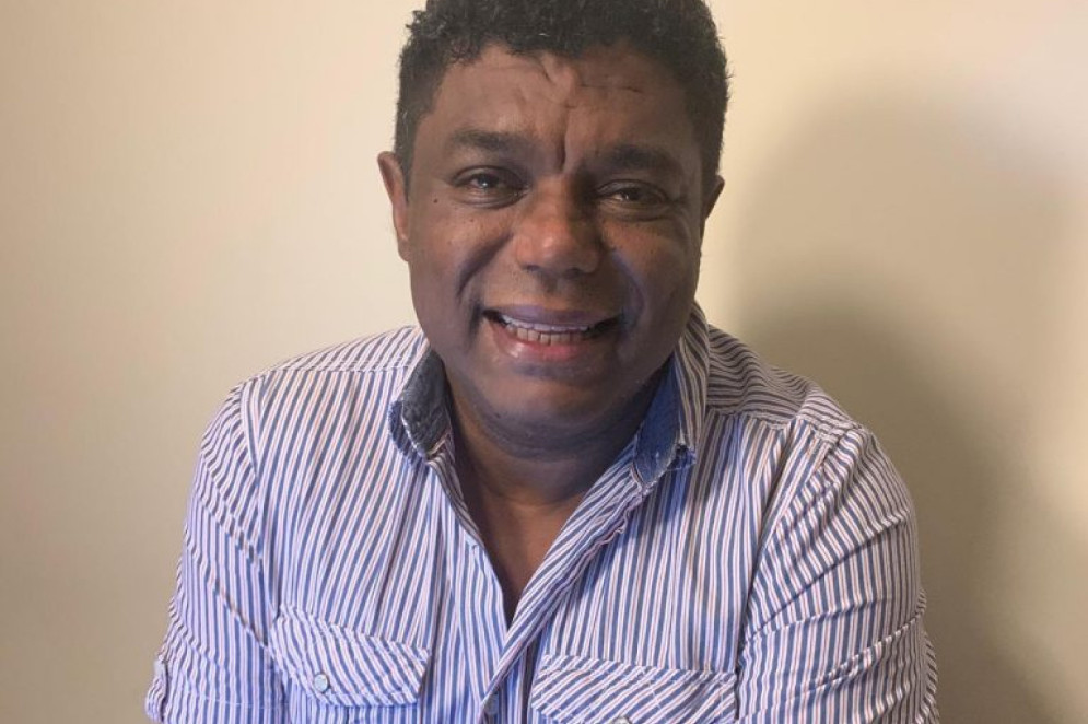 ParaTodosVerem: Fotografia do professor Melgaço Júnior, da UniRio, um homem de pele escura e cabelo curto e cacheado sorri levemente em frente a uma parede de cor clara. Ele veste uma camisa social listrada em azul e branco, com o colarinho levemente abert(Foto: Arquivo Pessoal)