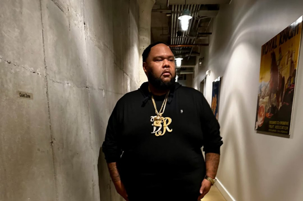ParaTodosVerem: A foto mostra o rapper Big Rich, um homem negro, de corpo robusto e rosto arredondado com nariz marcante e lábios cheios. Tem cabelos e barba curtos e escuros. Veste um moletom preto com um pingente dourado em formato da letra "R", várias correntes douradas no pescoço, calça escura e um relógio no pulso. Sua expressão é neutra e a postura ereta, em um ambiente casual. O visual mistura estilo casual com joias chamativas(Foto: Reprodução / Instagram )