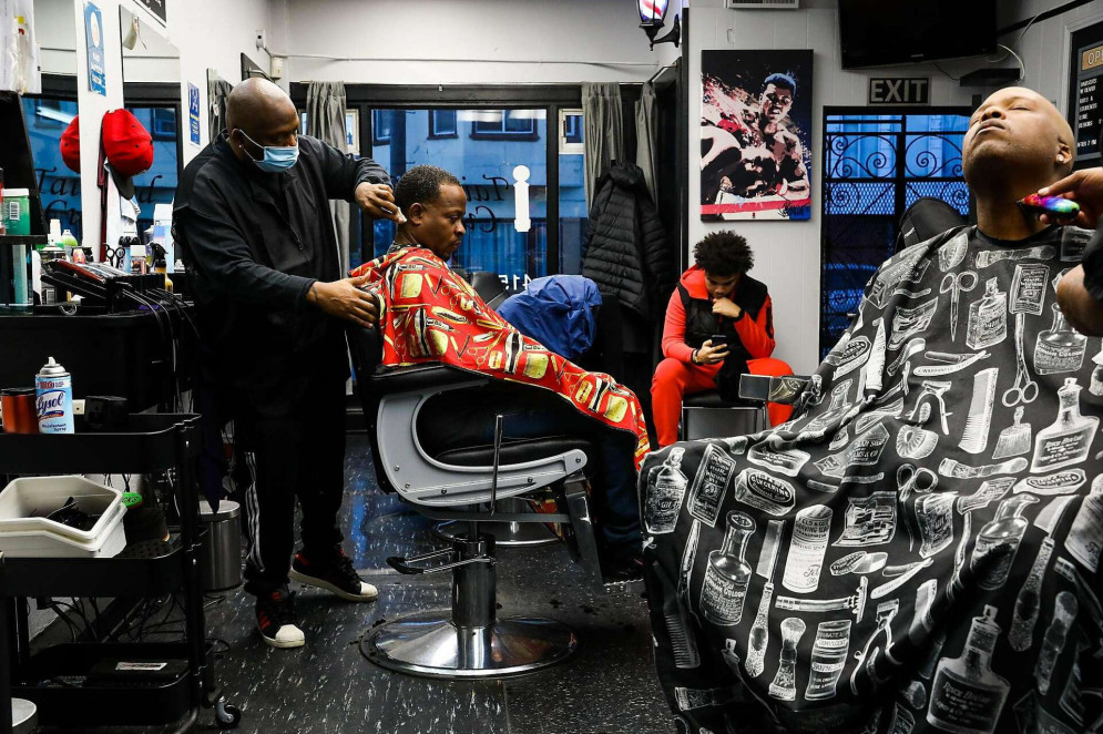 ParaTodosVerem: Foto da barbearia Tailored Cuts, em São Francisco nos EUA, à esquerda, um barbeiro de máscara corta o cabelo de um cliente sentado em uma cadeira giratória, coberto por uma capa vermelha com estampas de produtos de barbearia. À direita, outro cliente está reclinado, recebendo acabamento no pescoço com uma máquina elétrica, protegido por uma capa preta com desenhos de utensílios e produtos de barbearia em branco. A barbearia tem paredes claras, chão escuro com detalhes claros e pôsteres decorativos, incluindo uma imagem icônica do boxeador Muhammad Ali(Foto: Yalonda M. James / Tailored Cuts)