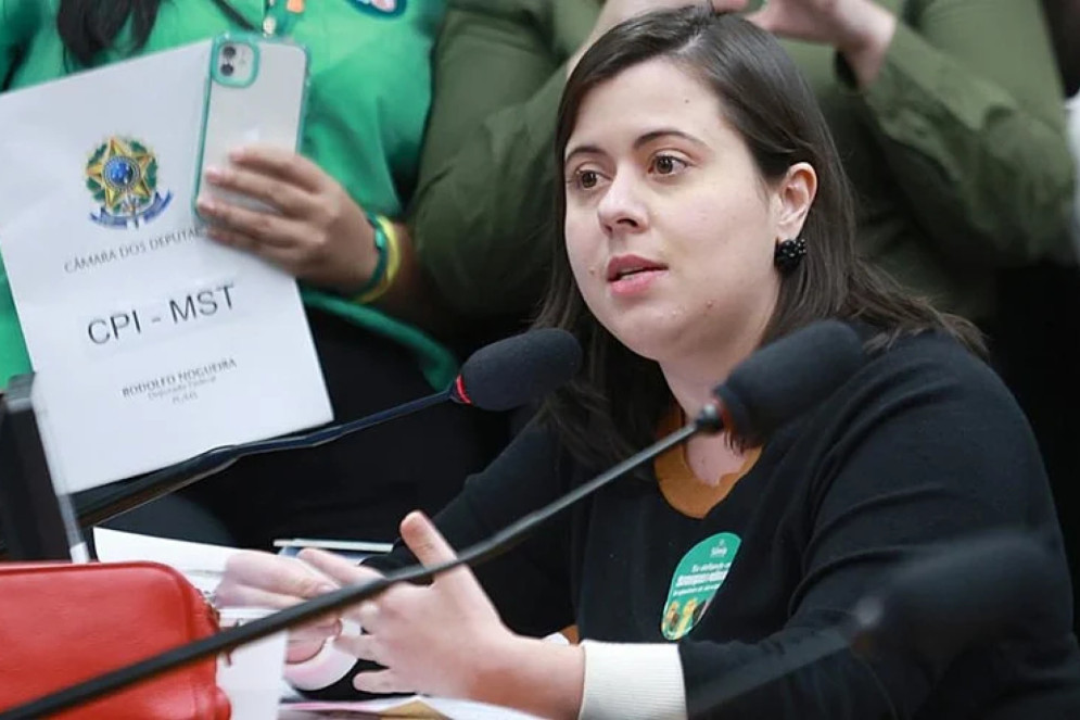ParaTodosVerem: Sâmia Bomfim, mulher branca de cabelos pretos, na CPI do MST em 2023. Ela fala no microfone, enquanto uma folha com o título CPI está ao fundo (Foto: Myke Sena/Câmara dos Deputados)