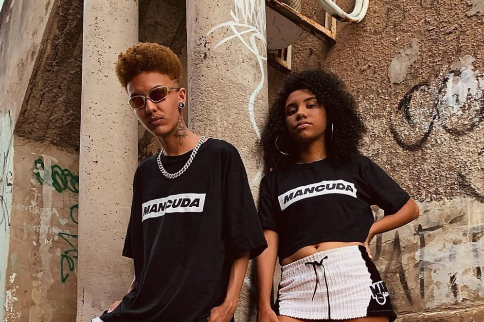 ParaTodosVerem: Em um ambiente urbano com um pilar de concreto e uma parede pichada ao fundo, os idealizadores da Mancuda posam, em pé. Ambos vestem roupas da marca. Carll, à esquerda, tem cabelo loiro descolorido, usa óculos escuros, uma corrente prateada, camiseta preta e bermuda branca e preta. Ele está apoiado no pilar. Nair, à direita, tem cabelo preto, longo e cacheado, e veste uma camiseta preta curta e um shorts branco e preto.(Foto: Arquivo Pessoal)