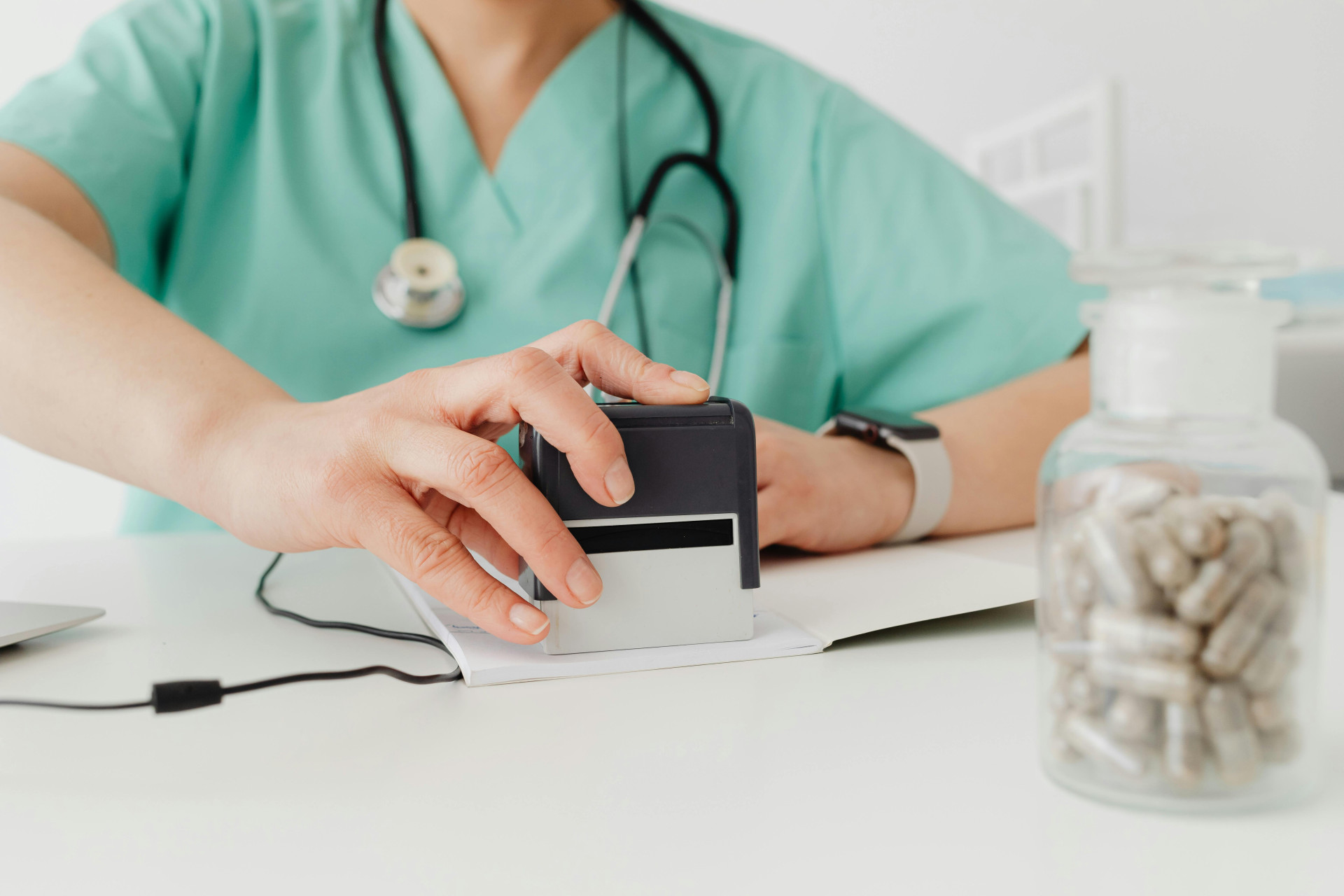 #ParaTodosVerem: pessoa com roupa médica verde e estetoscópio no pescoço carimba documentos sobre uma mesa. Em primeiro plano, há um frasco transparente com cápsulas (Foto: Pexels/Photo By: Kaboompics.com)