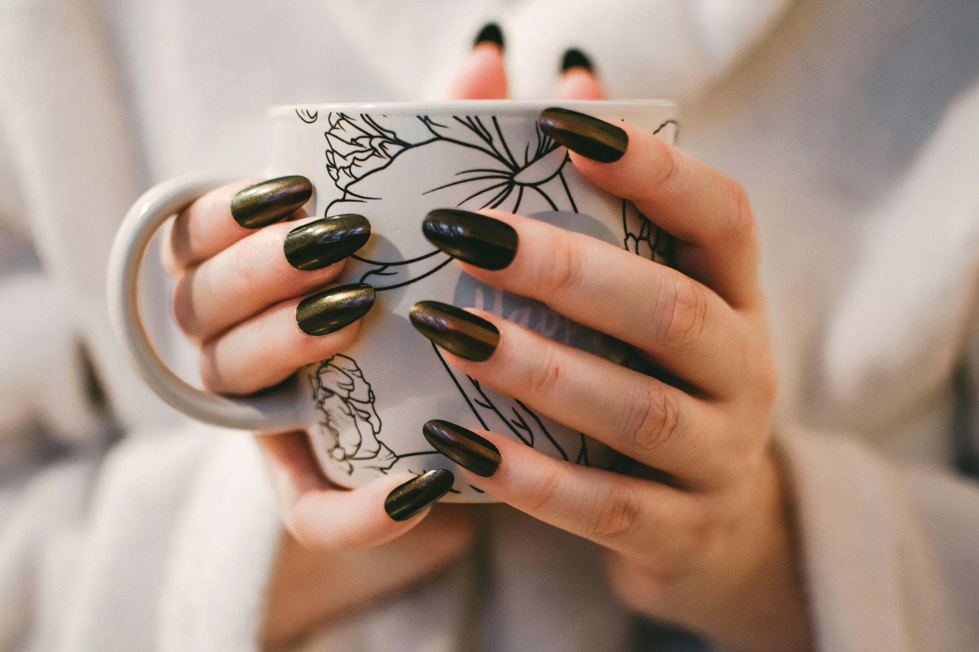 Mãos com unhas longas e esmaltadas em tom escuro seguram uma caneca branca com desenhos de flores em preto. Ao fundo, um tecido claro (Foto: Lisa from Pexels)