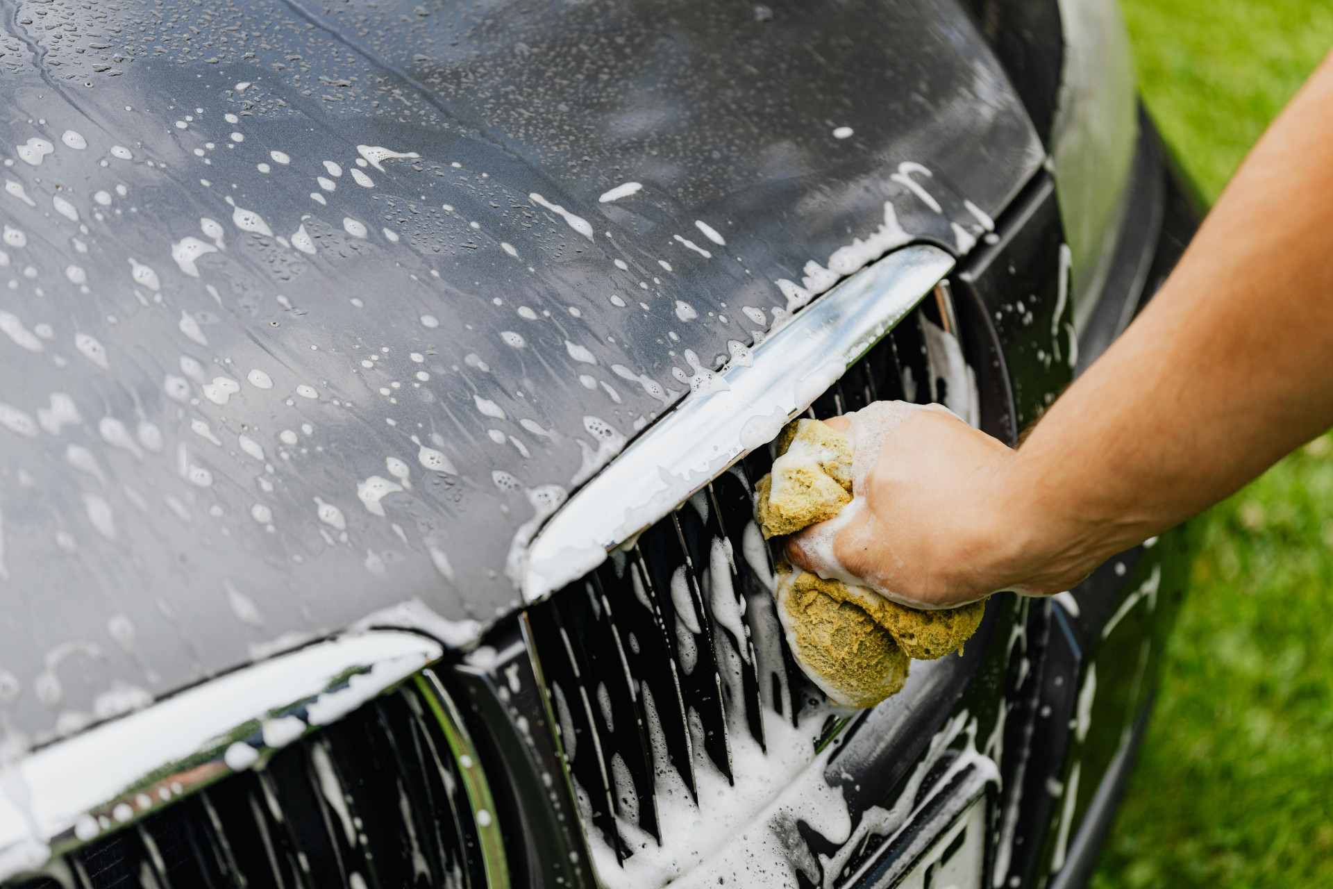 Uma mão esfrega a grade de um carro preto com uma esponja amarela cheia de espuma. O veículo está coberto de sabão, indicando que está sendo lavado ao ar livre (Foto: Pexels/Photo By: Kaboompics.com)
