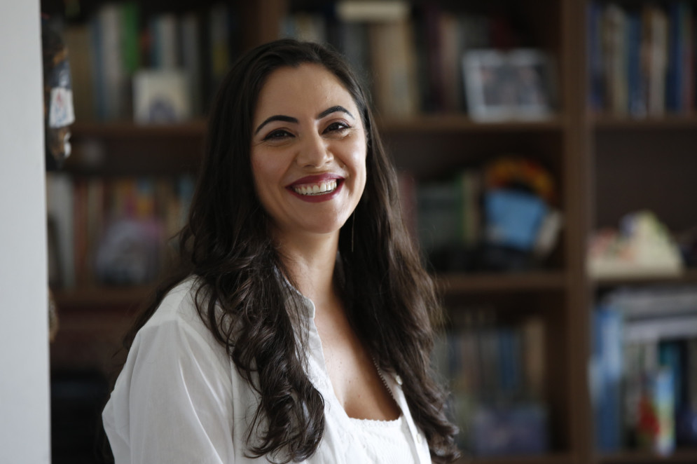 ParaTodosVerem: Juliana Dal Piva, jornalista. É uma mulher de cabelos escuros e longos, usa roupas claras. No fundo, estantes de livros(Foto: Daniel Marenco/Reprodução/UFSC)