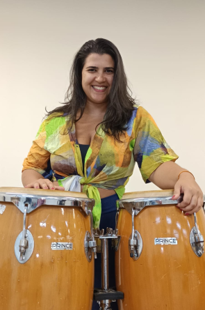 Catherine Furtado é professora do curso de Música da UFC e pesquisadora na área da percussão popular brasileira e africana e etnomusicologia. Está em pós-doutorado na África e é autora do livro "No solar dos ritmos". Na imagem ela aparece sorrindo apoiada em dois tambores de batuque(Foto: Catherine Furtado/Acervo pessoal)